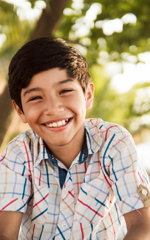 A portrait of a cheerful young boy with a beaming smile, enjoying a sunny day outdoors with a blurred background.